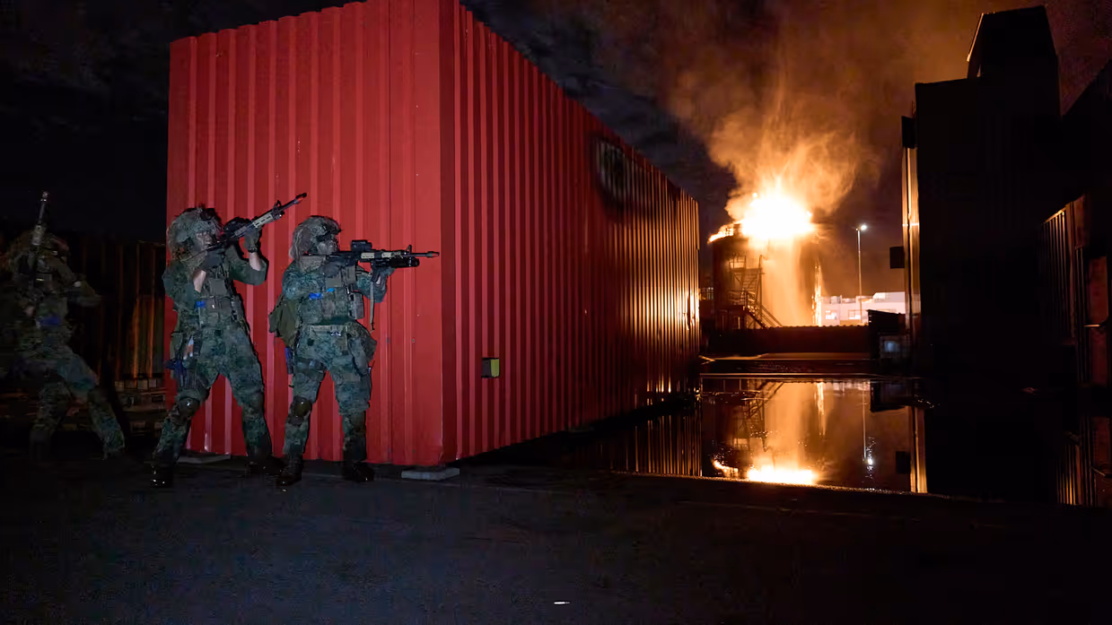 3 mariniers schuilen achter een container terwijl er om de hoek brand lijkt te woeden op een verhoogde installatie.