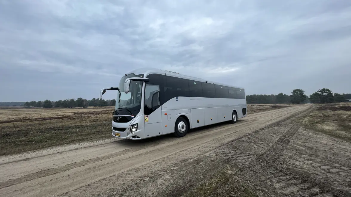 Een grote, witte touringcar rijdt over een onverhard, zanderig pad midden in een open, vlak landschap met lage struiken en een bewolkte lucht.