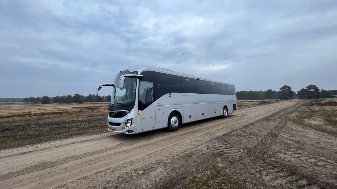 Een grote, witte touringcar rijdt over een onverhard, zanderig pad midden in een open, vlak landschap met lage struiken en een bewolkte lucht.