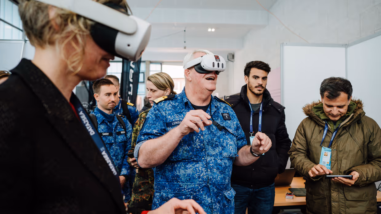 Een man in blauw uniform en een vrouw met VR-bril, anderen kijken toe in een lichte ruimte. 