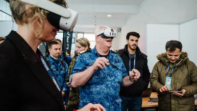 Een man in blauw uniform en een vrouw met VR-bril, anderen kijken toe in een lichte ruimte. 