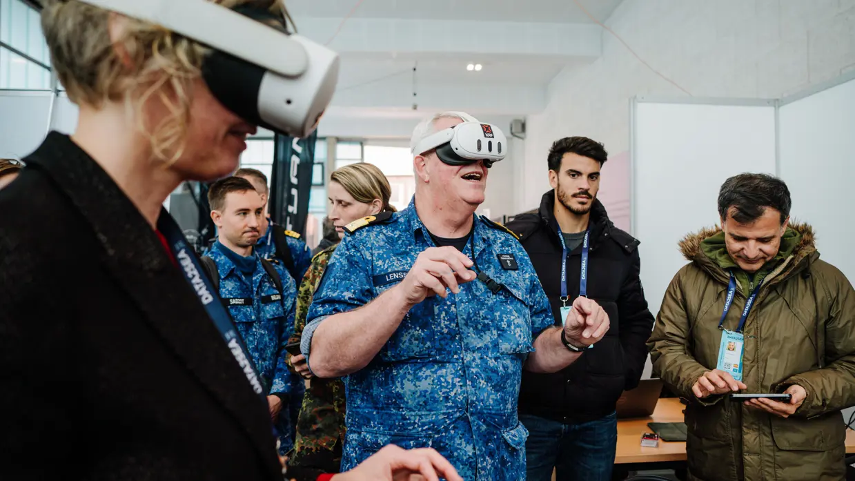 Een man in blauw uniform en een vrouw met VR-bril, anderen kijken toe in een lichte ruimte. 