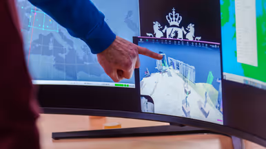 Groep mensen staat rond stand op conferentievloer. Een man wijst op computerscherm in een stand op de conferentie en een man in blauwe trui op de rug gezien zit tussen 2 mannen met VR-bril op.