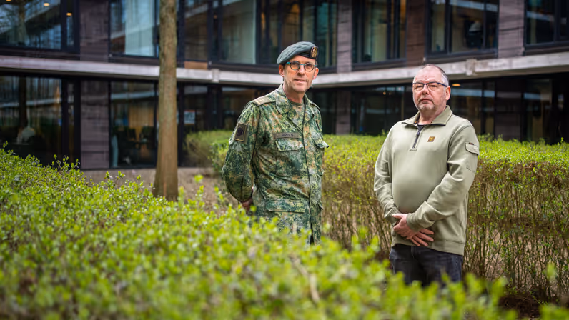 Militair en burgermedewerker Defensie staan naast elkaar achter een heg bij een kantoorgebouw.