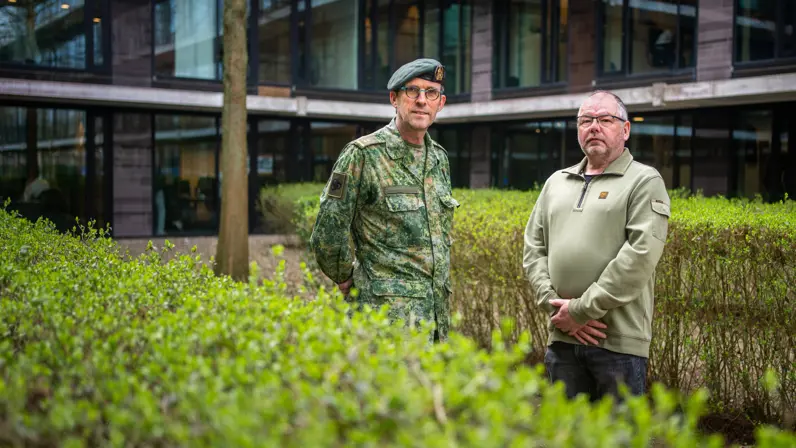 Militair en burgermedewerker Defensie staan naast elkaar achter een heg bij een kantoorgebouw.