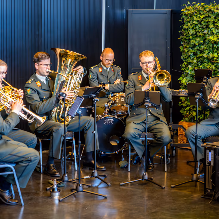 Een zestal militairen in donkergroen uniform speelt muziek. De militairen maken daarbij gebruik van blaasinstrumenten en een keybord. 
