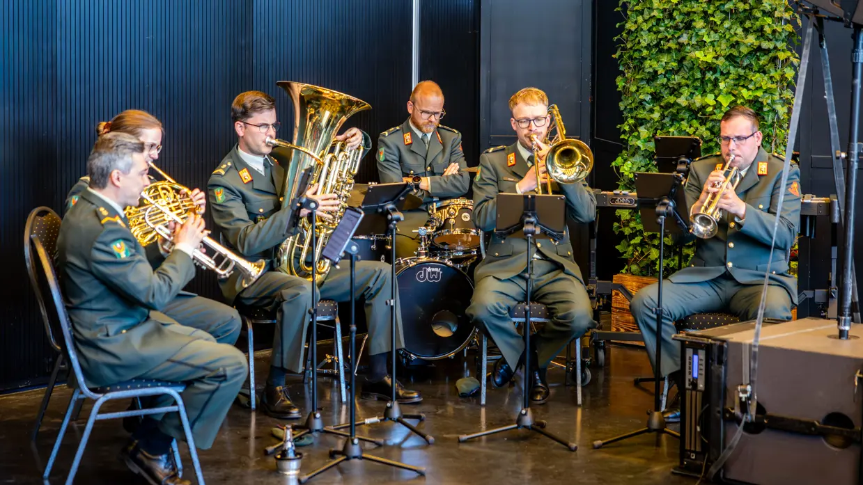 Een zestal militairen in donkergroen uniform speelt muziek. De militairen maken daarbij gebruik van blaasinstrumenten en een keybord. 