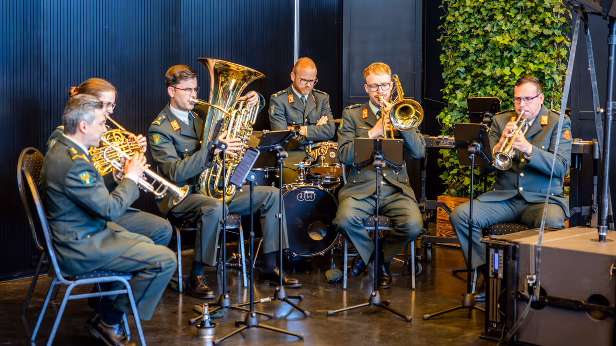 Een zestal militairen in donkergroen uniform speelt muziek. De militairen maken daarbij gebruik van blaasinstrumenten en een keybord. 