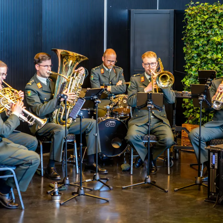 Een zestal militairen in donkergroen uniform speelt muziek. De militairen maken daarbij gebruik van blaasinstrumenten en een keybord. 