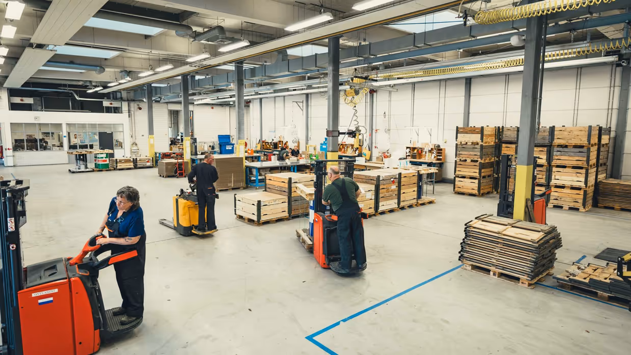 Overzicht van de grijze loods. Er zijn diverse lange tafels, boxpallets en heftrucks te zien. 