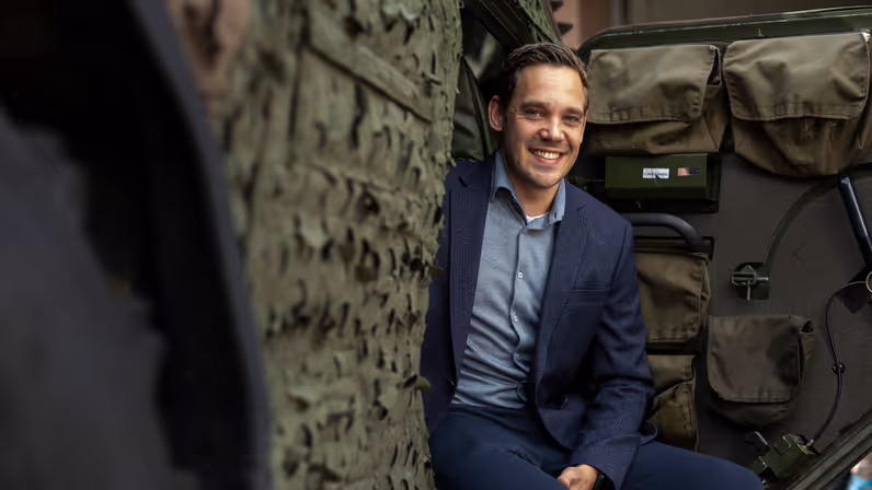 Een lachende man in blauw pak zit op een militair voertuig. 