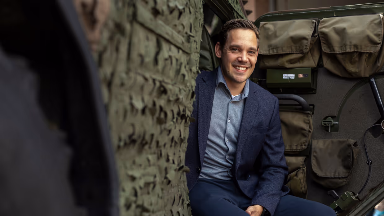 Een lachende man in blauw pak zit op een militair voertuig. 