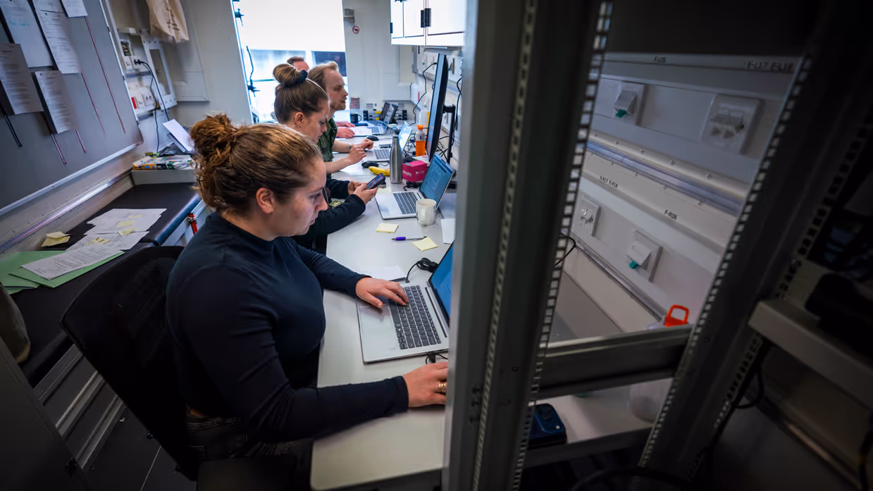 Twee vrouwen en twee mannen, waarvan één in uniform, zitten achter computers in een conrtainer.