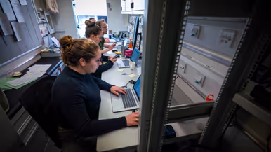 Twee vrouwen en twee mannen, waarvan één in uniform, zitten achter computers in een conrtainer.