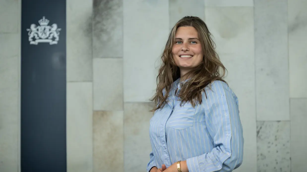 Vrouw met lang donkerblond haar en lichtblauw krijtjesoverhemd aan poseert met handen over elkaar.