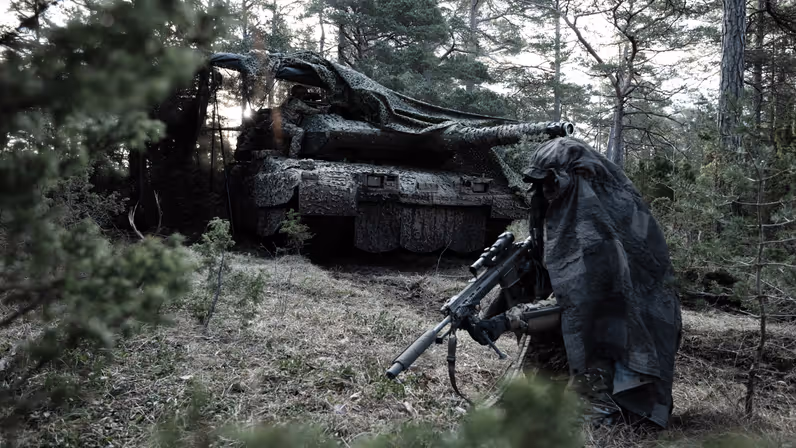 Een soldaat en een gepantserd militair voertuig in het bos, beiden gecamoufleerd.