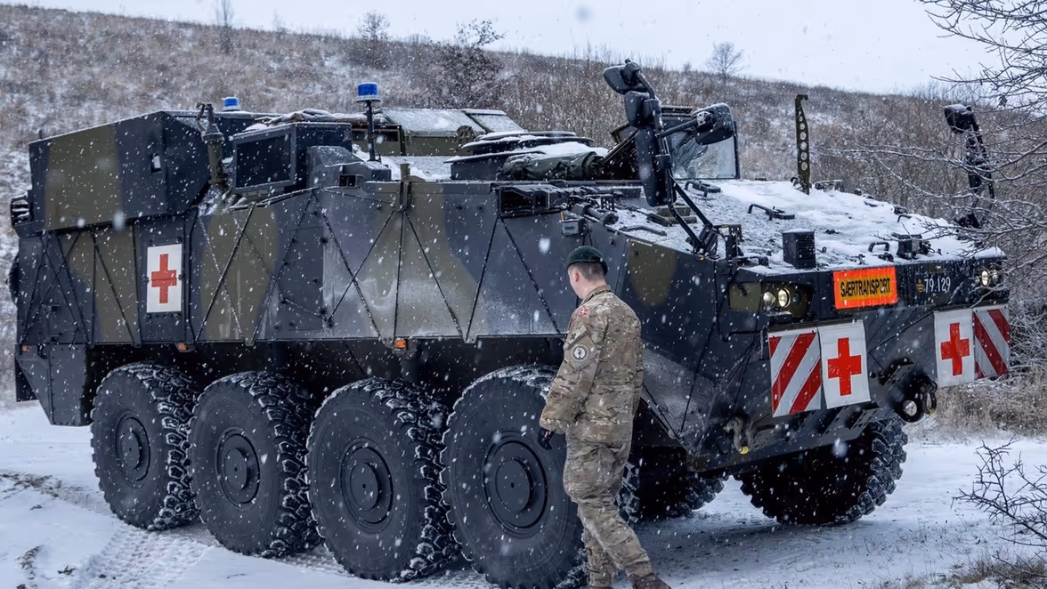 Een militair in winterkleding loopt naast een groot, zwaar pantservoertuig met rode kruizen en sneeuw op de weg en een ander voertuig, tijdens een sneeuwbui. 