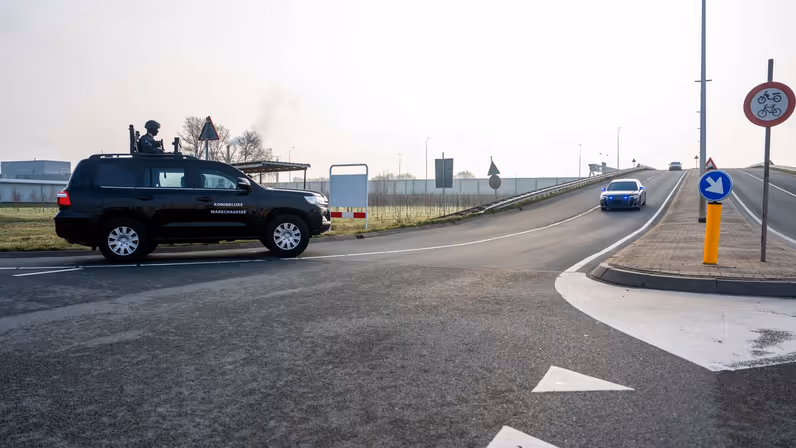 Een bochtige weg met een zwart politievoertuig van de Koninklijke Marechaussee, een auto op de weg, en verkeersborden langs een brede, rustige asfaltweg.