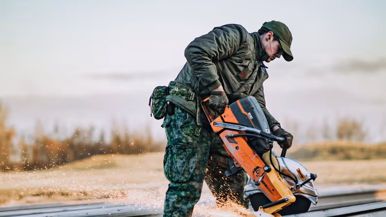 Een militair zaagt met een vonkende zaag een metaalrail buiten in de natuur. 