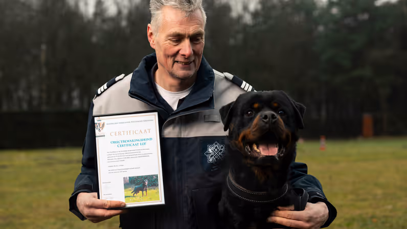 +alttekst: Man, geknield in uniform, houdt trots een certificaat vast. Naast hem zit een rood-zwarte Rottweiler met uitstekende tong in een groene, bosrijke omgeving. 