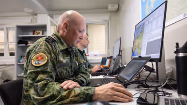 Links: Een militair werkt geconcentreerd achter een bureau met computerschermen. Op de schermen zijn kaarten te zien, terwijl een collega op de achtergrond ook bezig is. Rechts: Een close-up van een militair uniform met een rond embleem ‘Casualty Move 2026’ en een rood kruis. Op de achtergrond is wazig een persoon in oranje zichtbaar.