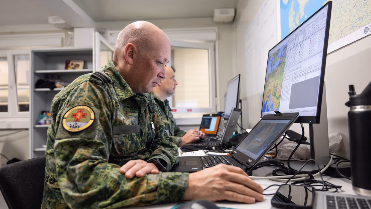 Links: Een militair werkt geconcentreerd achter een bureau met computerschermen. Op de schermen zijn kaarten te zien, terwijl een collega op de achtergrond ook bezig is. Rechts: Een close-up van een militair uniform met een rond embleem ‘Casualty Move 2026’ en een rood kruis. Op de achtergrond is wazig een persoon in oranje zichtbaar.