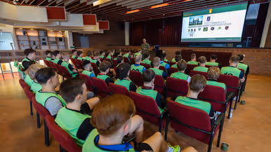 Een groep jongeren in gele vesten luistert in een zaal naar een militair die een presentatie geeft over organisatie en verschillende logo’s op een groot scherm.