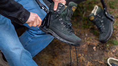 Iemand trekt een groene bergschoen aan met een zwarte beugel, buiten op een bosbodem met bladeren.