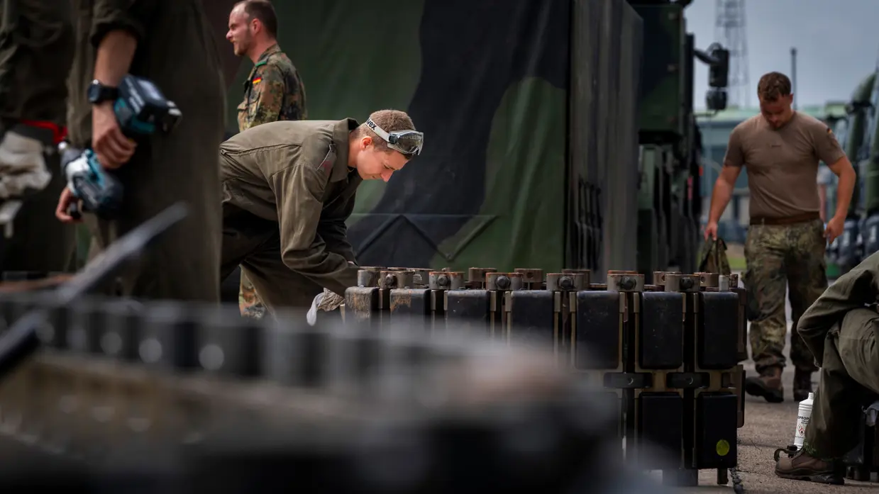 Soldaten in uniform werken met zware metalen onderdelen bij een groene militaire vrachtwagen, omringd door gereedschap en uitrusting.