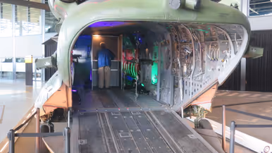 Inkijk in een grote, openstaande militaire helikopter; binnenin zie je stoelen, apparatuur en knipperende groene lichten, terwijl iemand binnen staat.