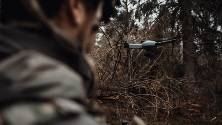 Een grijze drone vliegt voor een militair, die vanaf de achterkant wordt bezien 