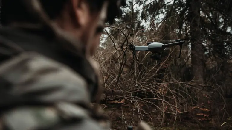 Een grijze drone vliegt voor een militair, die vanaf de achterkant wordt bezien 