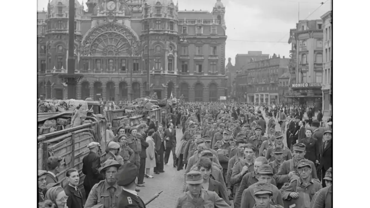 Een drukke straat met soldaten en burgers, een groot station met klok en klassieke architectuur op de achtergrond.