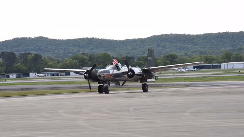 Een oud militair vliegtuig met twee propellers staat op een startbaan, omringd door groene heuvels en gebouwen, klaar om te taxiën.