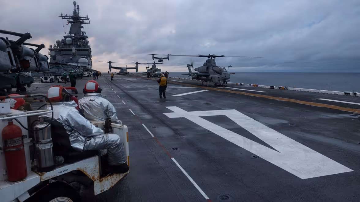 een druk vliegdek met helikopters en vliegtuigen. Twee bemanningsleden in witte pakken zitten op een voertuig, terwijl anderen op de achtergrond lopen. Zee en wolkenlucht omringen het schip.