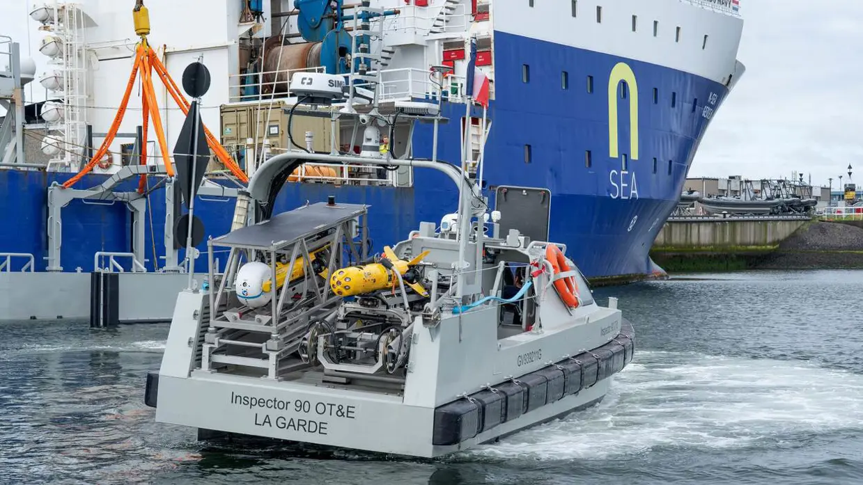 Een grijs bootje met achterop verschillende drones. Dit vaart langs een groot vrachtschip.