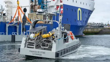 Een grijs bootje met achterop verschillende drones. Dit vaart langs een groot vrachtschip.