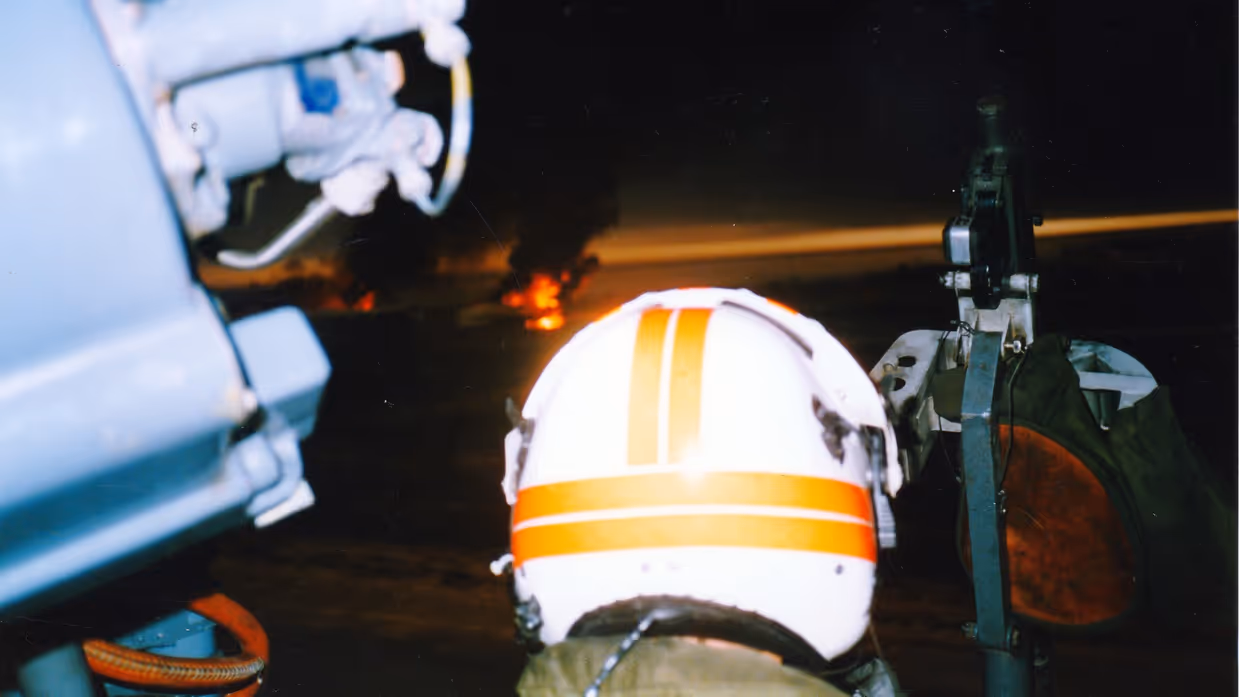 Een bemanningslid van een helikopter, met een witte helm met oranje strepen, kijkt in een donkere lucht met vuur en rook op de achtergrond.