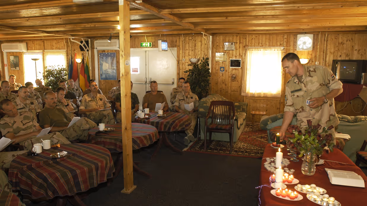 Militairen in uniform zitten in een houten ruimte; één man steekt kaarsen aan op een tafel met bloemen, boeken en theelichtjes. 