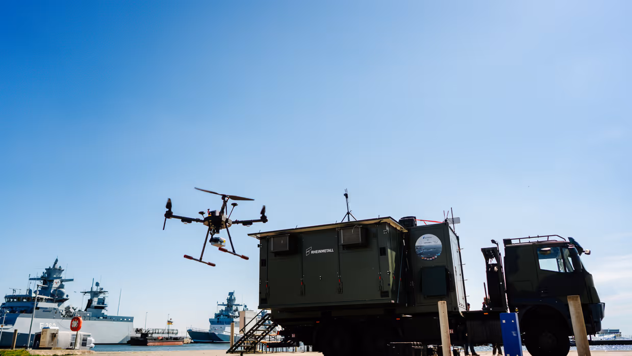 Een drone vliegt boven een groene vrachtwagen bij een haven met schepen op een zonnige dag. 