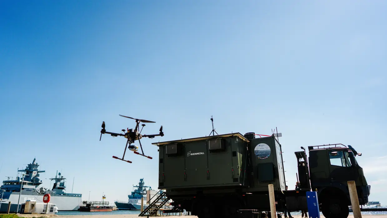 Een drone vliegt boven een groene vrachtwagen bij een haven met schepen op een zonnige dag. 
