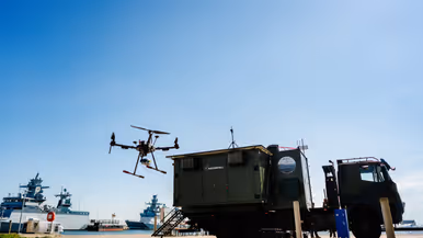 Een drone vliegt boven een groene vrachtwagen bij een haven met schepen op een zonnige dag. 