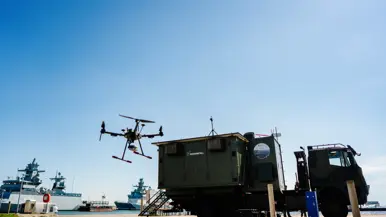 Een drone vliegt boven een groene vrachtwagen bij een haven met schepen op een zonnige dag. 
