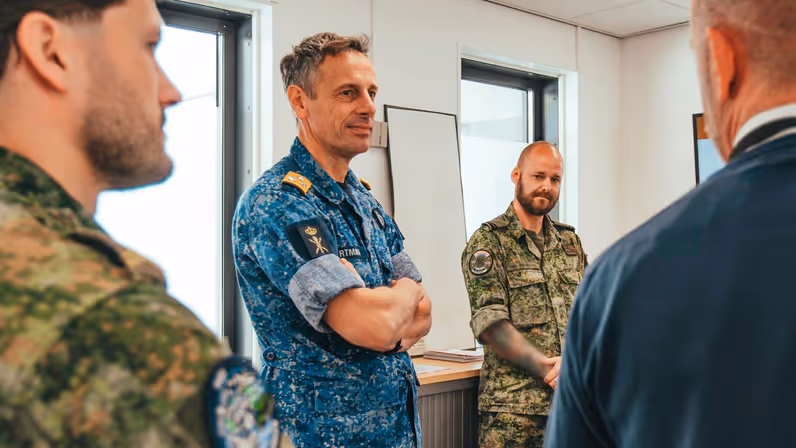 Vice-admiraal Jan Willem Hartman, in blauw gevechtstenue, staat tussen twee militairen in groen uniform. Hij luistert naar een derde persoon in net pak.