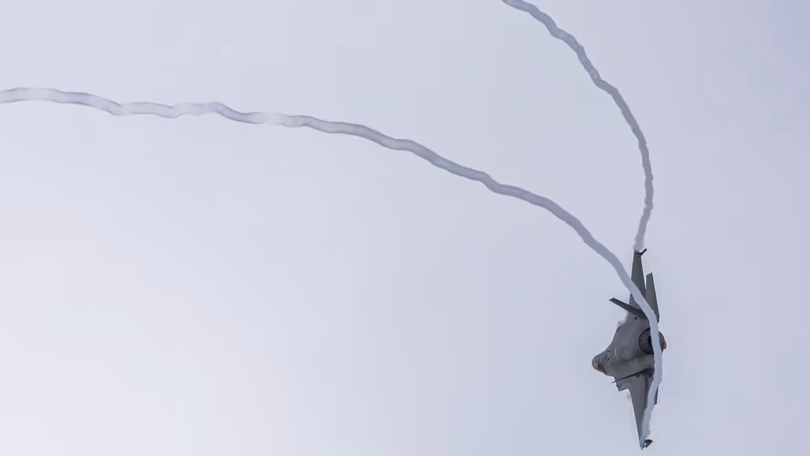 Een straaljager vliegt ondersteboven hoog in de lucht en trekt een witte rooklijn achter zich aan, tegen een lichte, heldere hemel.    