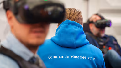 Groep mensen staat rond stand op conferentievloer. Een man wijst op computerscherm in een stand op de conferentie en een man in blauwe trui op de rug gezien zit tussen 2 mannen met VR-bril op.