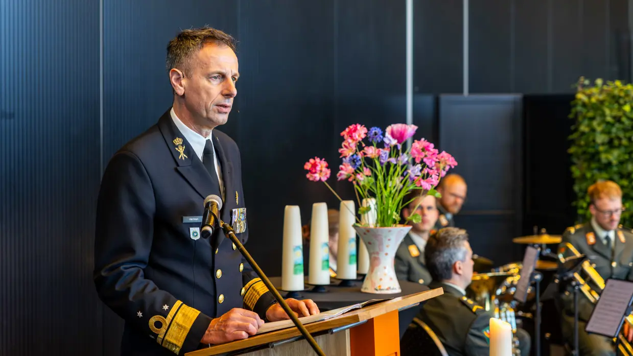 Vice-admiraal Jan Willem Hartman spreekt de aanwezigen toe. Dat doet hij via een microfoon. Naast hem staat een vaas met gekleurde bloemen. 