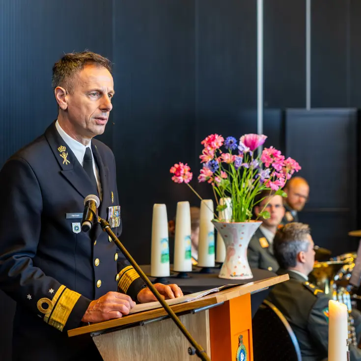Vice-admiraal Jan Willem Hartman spreekt de aanwezigen toe. Dat doet hij via een microfoon. Naast hem staat een vaas met gekleurde bloemen. 