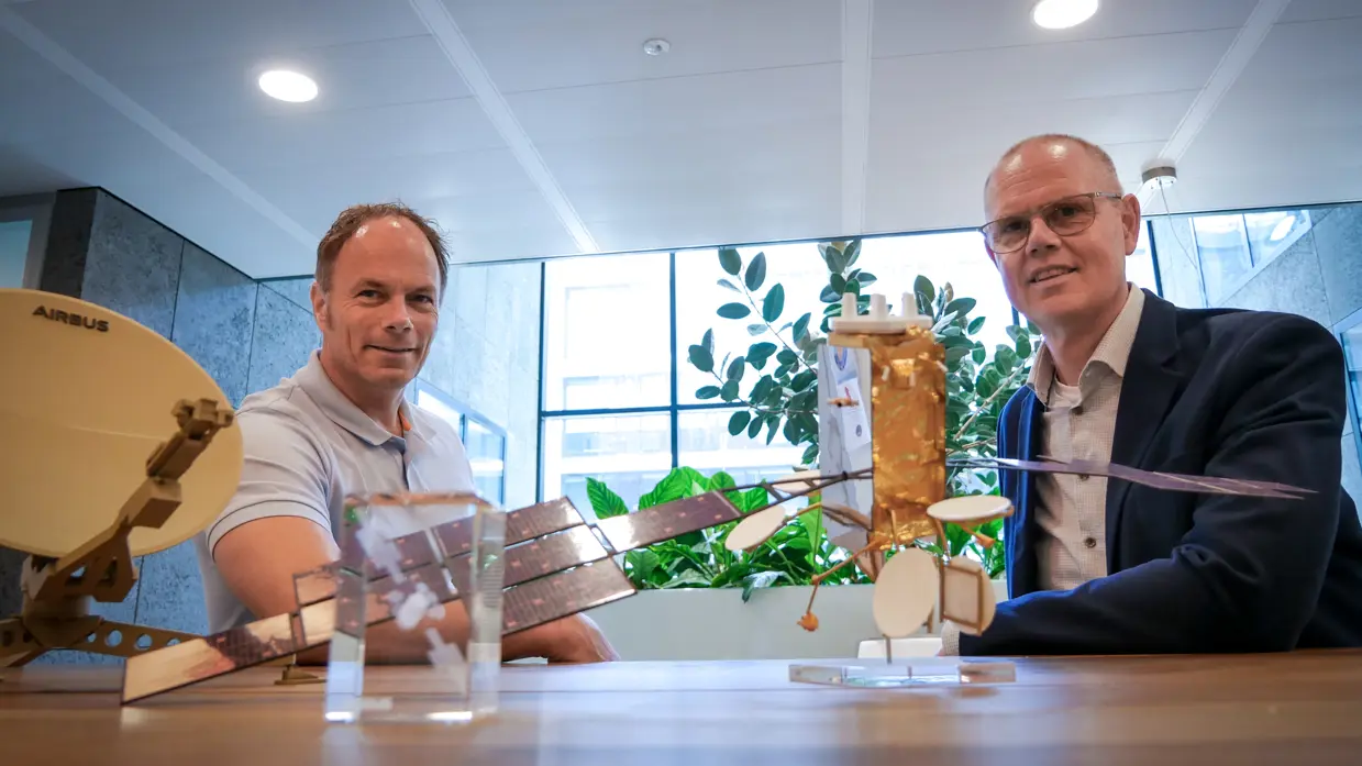 Martijn en Gerben poseren naast elkaar, zittend aan een tafel. Voor hen is een miniatuursatelliet opgesteld.
