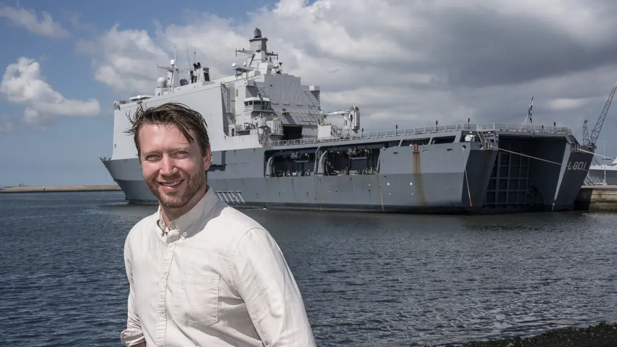 Een man in witte blouse staat op de kade van de marinebasis in Den Helder. Achter hem ligt een groot, grijs marineschip.